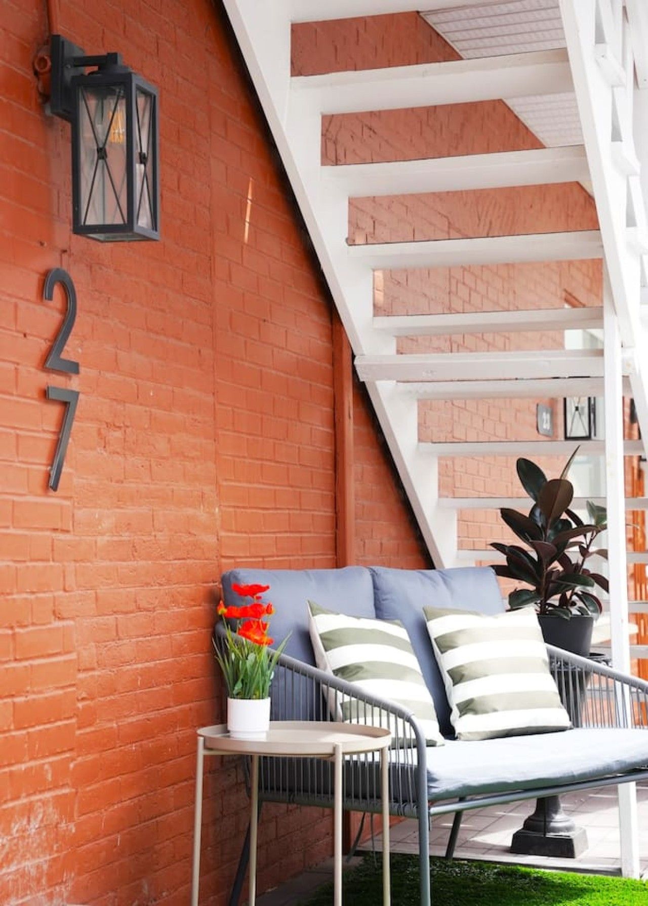 A brick wall with a number sign, a white staircase, a metal bench with cushions, and a potted plant.