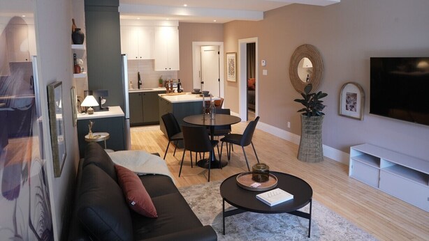 Un salon moderne avec un canapé noir, une table basse ronde et un téléviseur. On y trouve également un coin repas avec une table et des chaises, et une cuisine à l’arrière-plan.