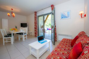 Ein Zimmer mit einem weißen Tisch, einem Laptop, einem Sofa, einem Fernseher und einem Balkon mit roten Vorhängen.