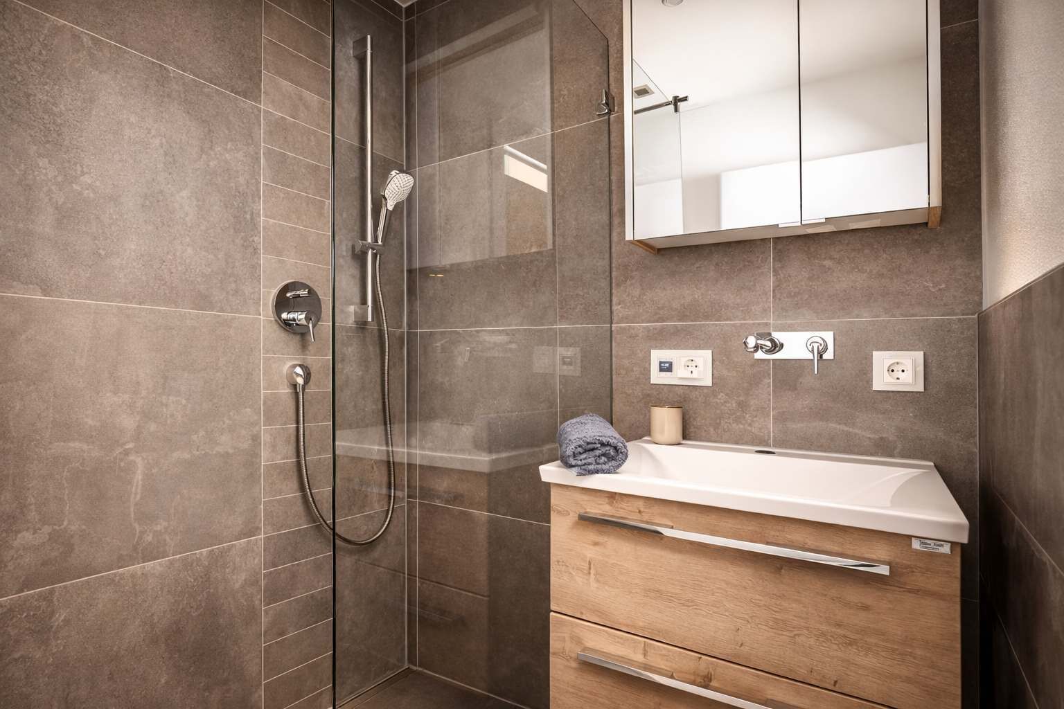 Un baño moderno con cabina de ducha de vidrio, tocador de madera con lavamanos blanco y un espejo sobre el tocador.