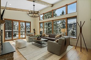 Ein geräumiges Wohnzimmer mit einer großen Couch, zwei Sesseln, einem Couchtisch und einem Kamin. Große Fenster bieten Blick auf schneebedeckte Bäume.