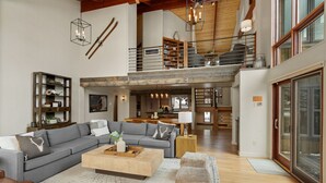 A spacious living area with a large sofa, a coffee table, and a fireplace. There is a staircase leading to an upper level with a balcony. - *New Listing* Swift Bear Ski-In/Out Lodge (Big Sky)