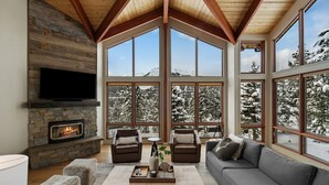 A cozy living room with a stone fireplace, large windows, and a view of snowy mountains. - *New Listing* Swift Bear Ski-In/Out Lodge (Big Sky)