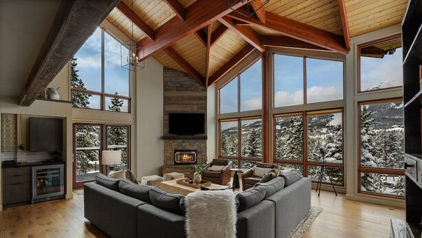 A spacious living room with a large sectional sofa, a fireplace, and large windows offering a view of snow-covered trees. - *New Listing* Swift Bear Ski-In/Out Lodge (Big Sky)