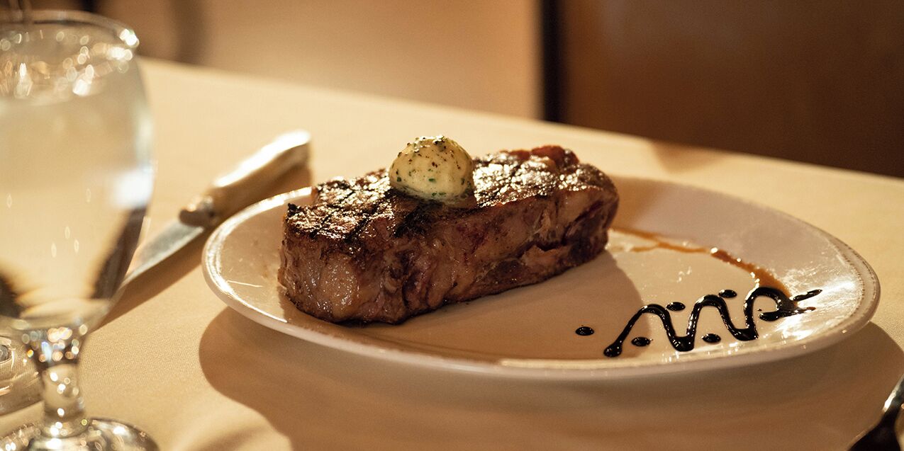 A steak with a sauce on a plate, a knife, and a glass of water on a table.