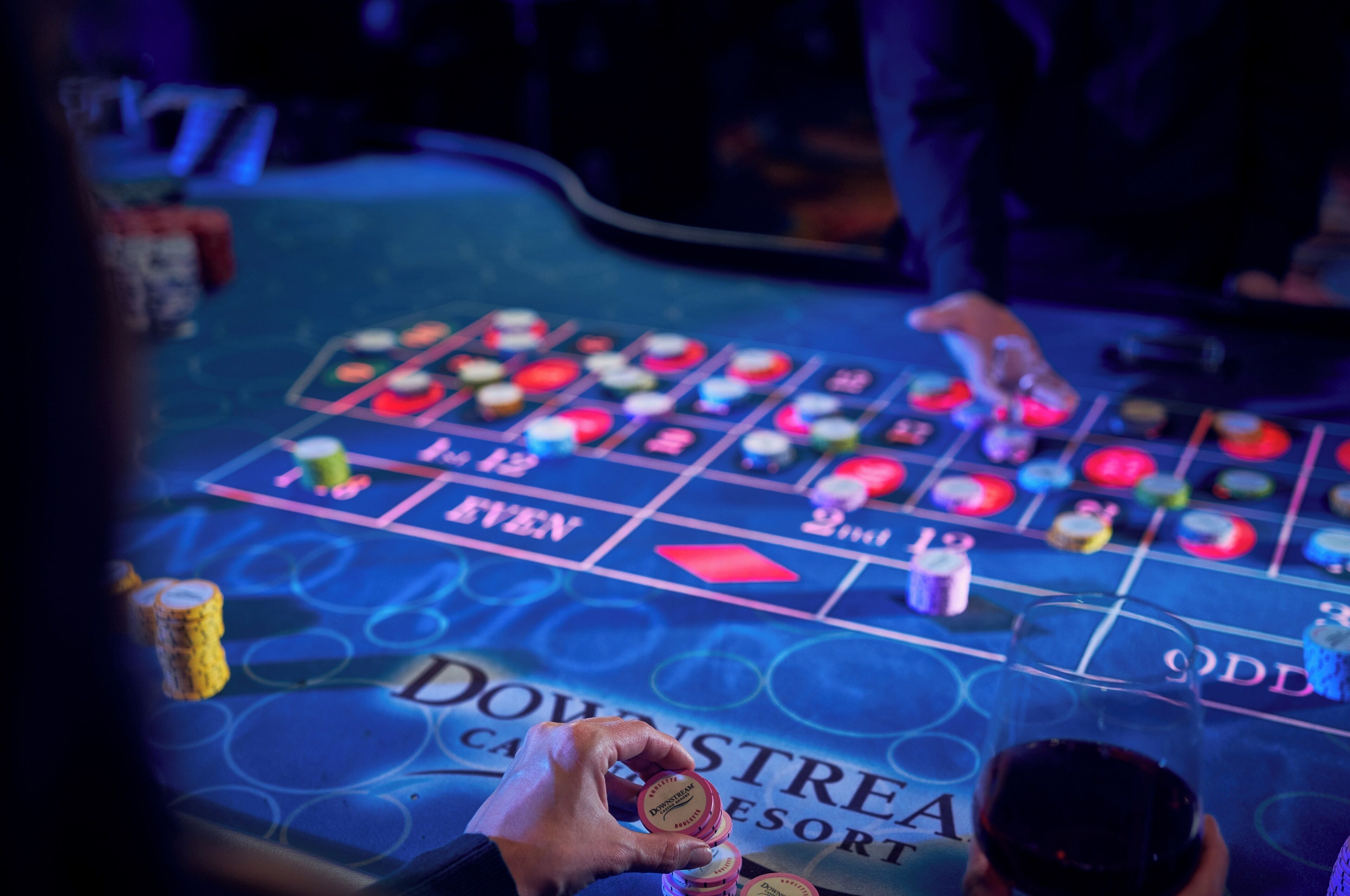 Ein Casinotisch mit Chips und einem Getränk, ein Blackjack-Tisch mit Karten und der Hand eines Dealers, sowie der Text 'DOWNTREAS' auf dem Tisch.