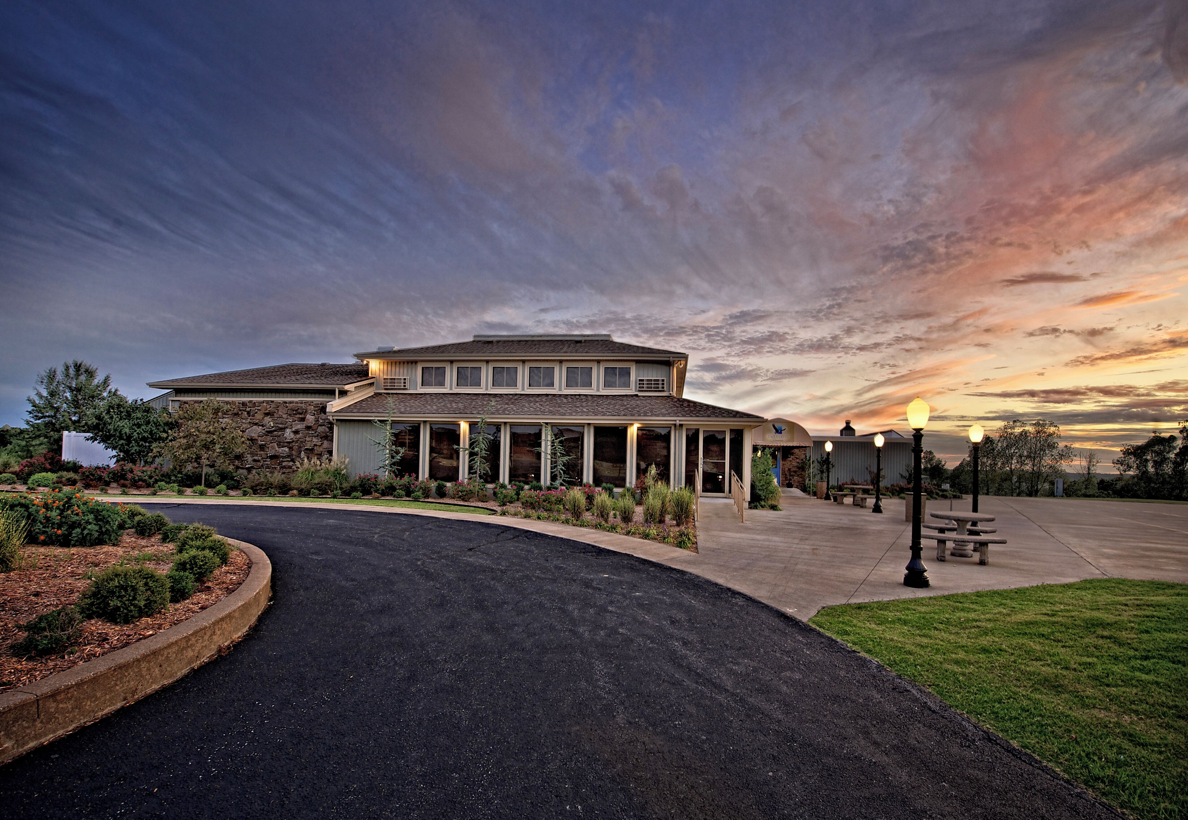 A building with a covered entrance, a paved driveway, and a landscaped area with shrubs and flowers.