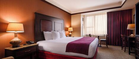 A hotel room with a large bed, two bedside tables with lamps, a desk with a chair, and a window with curtains.