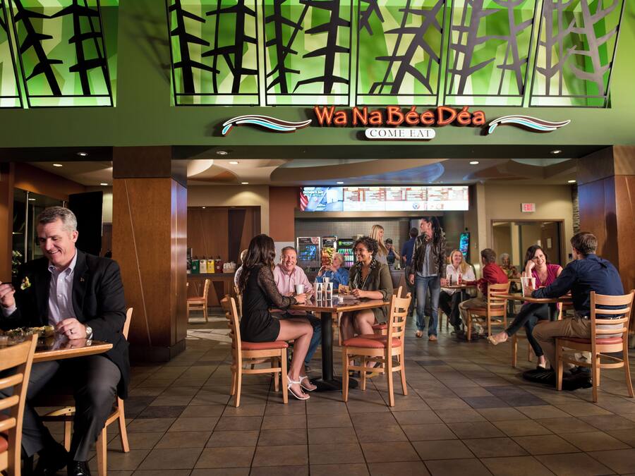 A restaurant interior with tables and chairs, a menu board, and a sign reading 'Wa Na Bee Déa Come Eat.'
