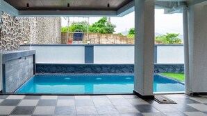 A covered swimming pool area with a stone wall, a pool with clear blue water, and a view of a green landscape. - Enjoy your staycation in lonavala with our luxurious villa. (Lonavala)