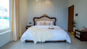 A bedroom with a large bed, a bedside table, a nightstand, a window with curtains, and a view of a pool. - Enjoy your staycation in lonavala with our luxurious villa. (Lonavala)
