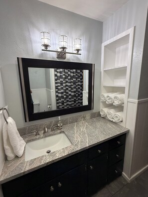 Une salle de bain dotée d’un comptoir en granit, d’un miroir à cadre noir, d’un pare-feu décoratif et d’un porte-serviettes.