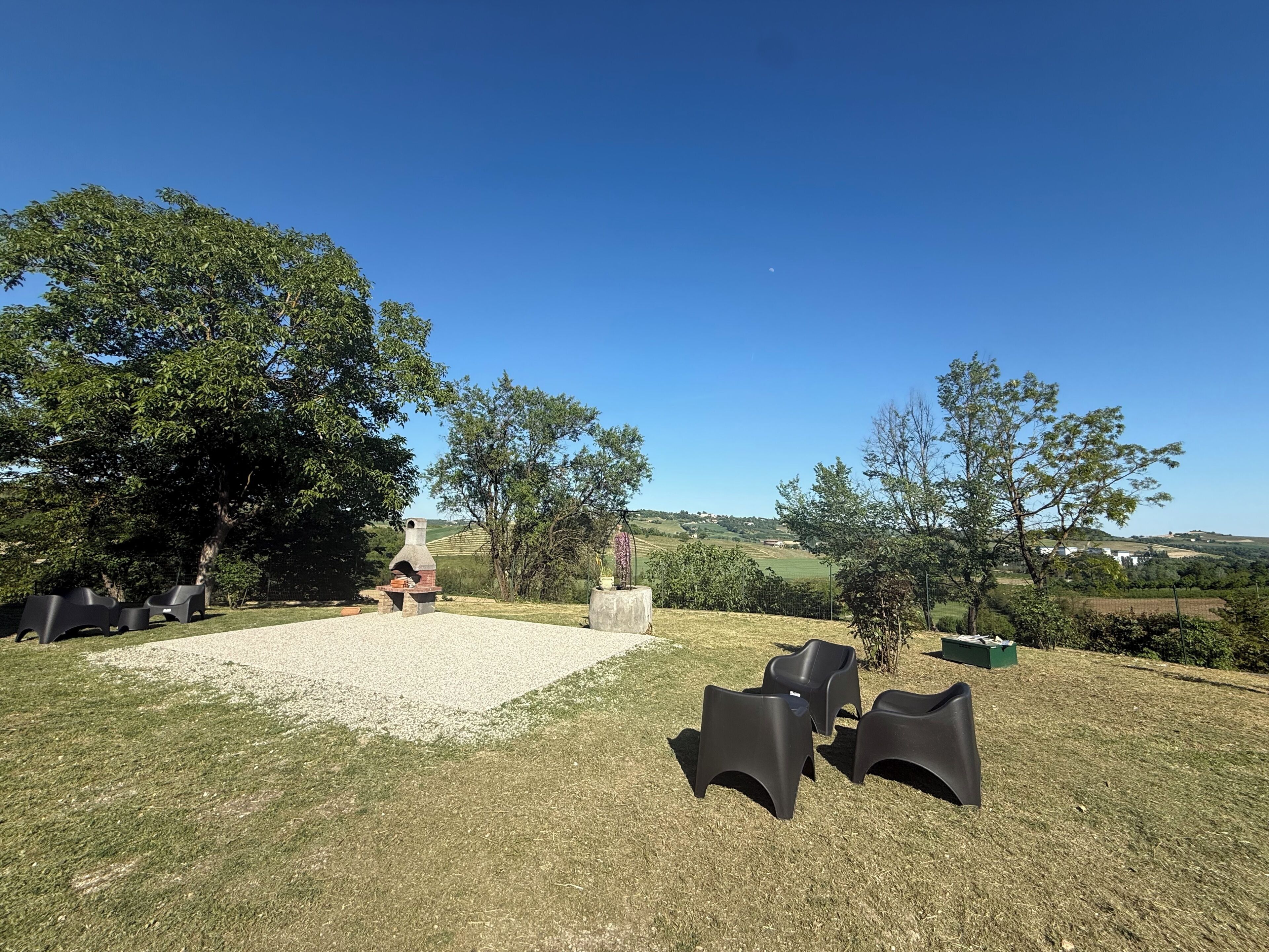 Un área de estar al aire libre con chimenea de piedra, rodeada de árboles y un cielo azul despejado.