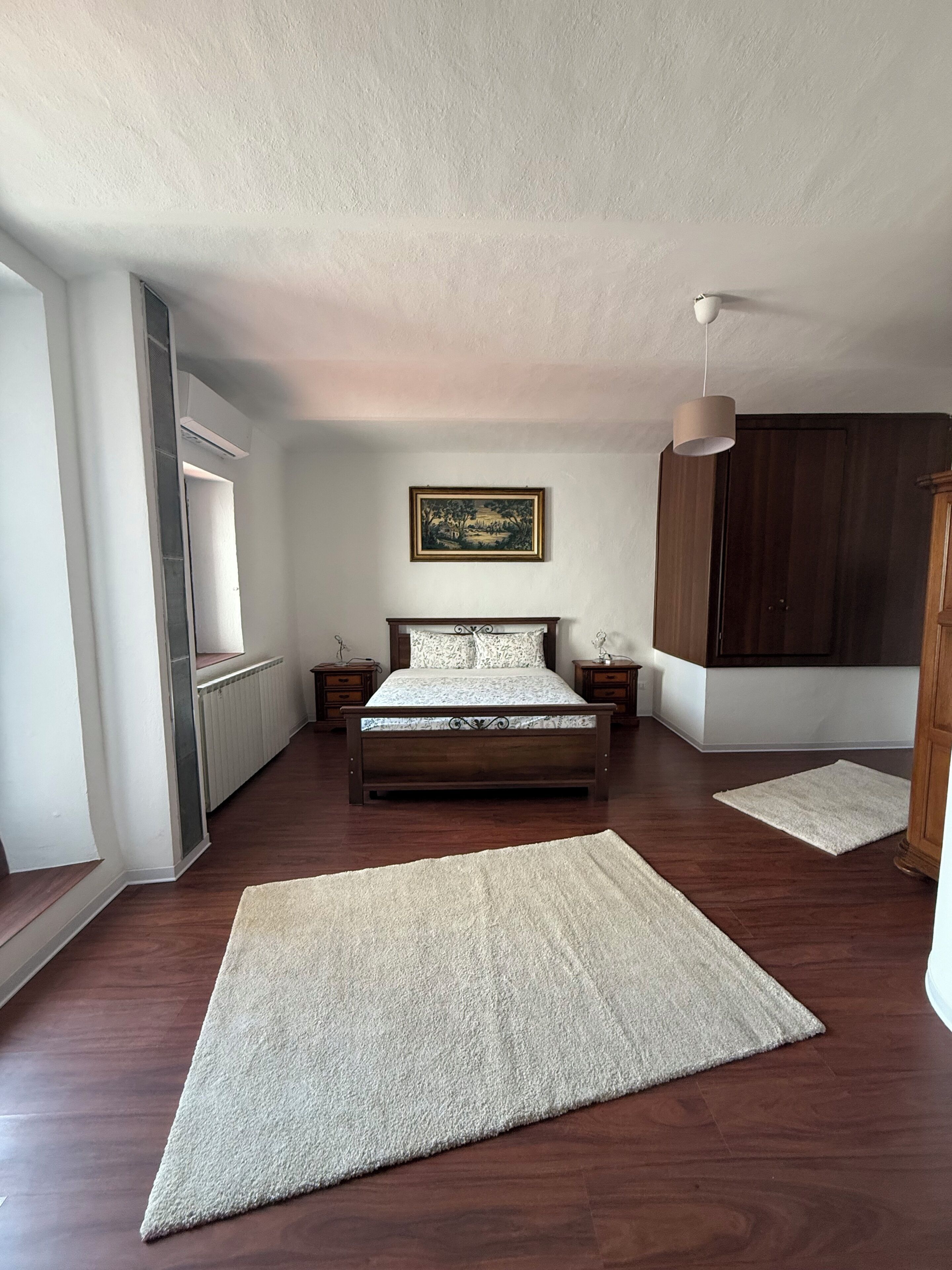 Quarto com cama, radiador, janela, quadro e guarda-roupa.