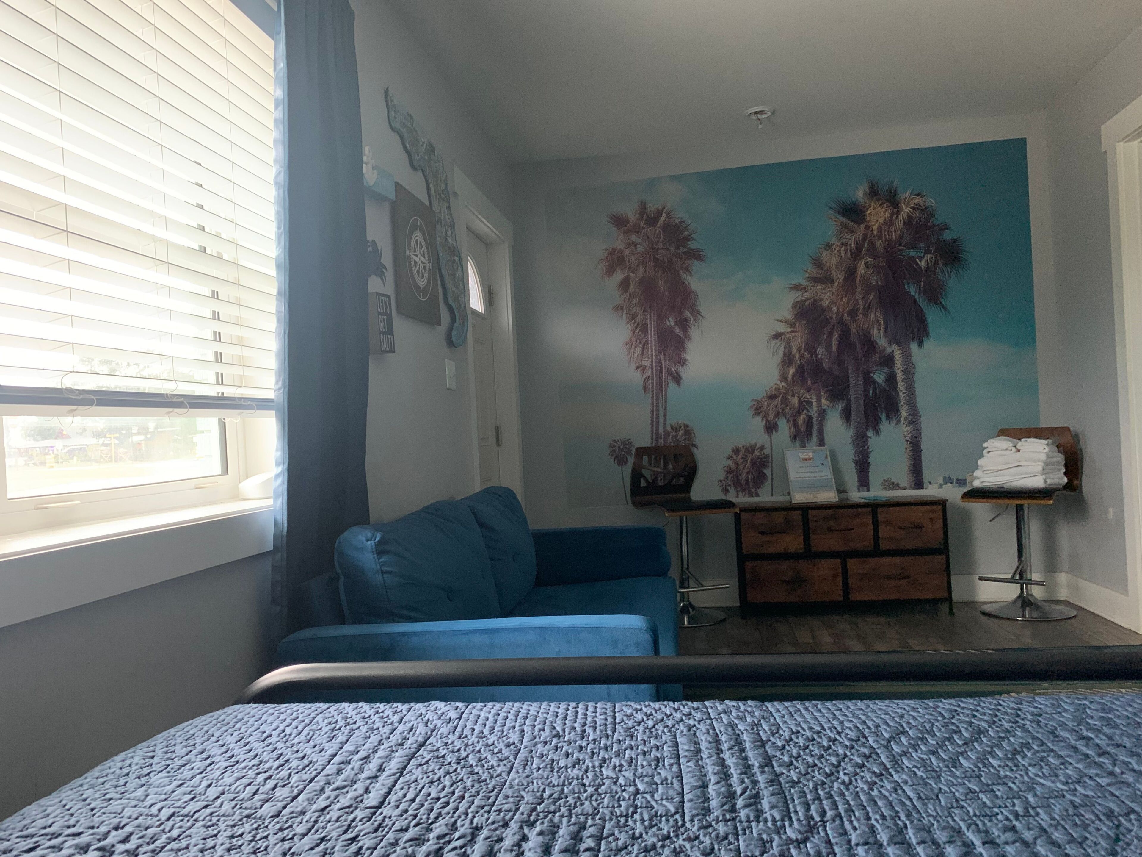 Une chambre avec un lit, un canapé bleu, une commode et une fresque murale représentant des palmiers.
