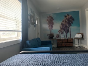 A bedroom with a bed, a blue sofa, a dresser, and a wall mural of palm trees.