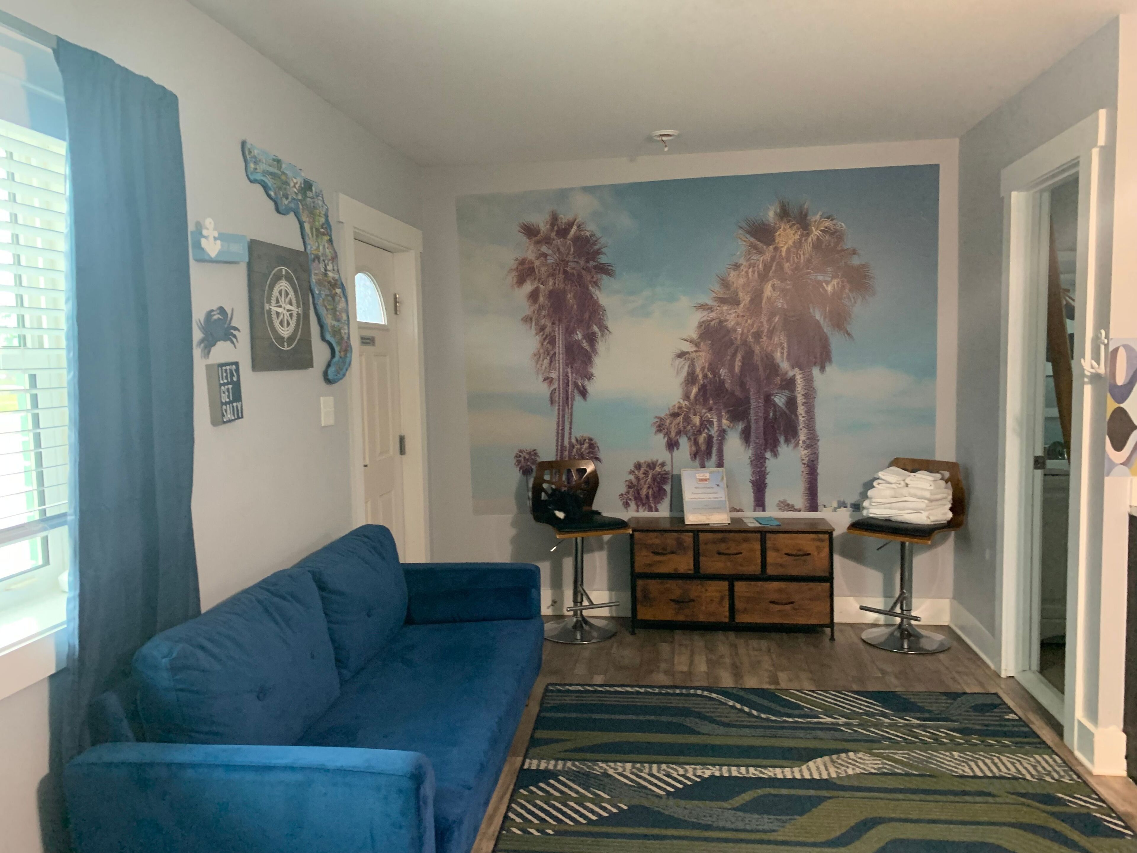 Ein Wohnzimmer mit blauem Sofa, einem gemusterten Teppich, einer Wandmalerei von Palmen, einem Holzschrank und einem kleinen Tisch mit Handtüchern.