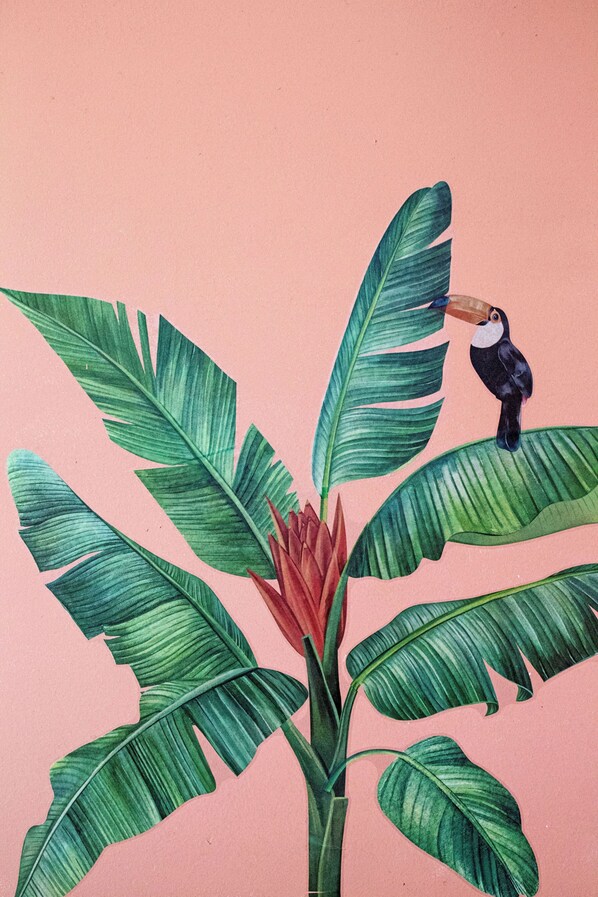 Tropical wall art featuring a banana plant and perched toucan against a pink background.
