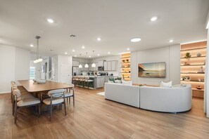 Eine moderne Küche mit weißen Schränken, ein Essbereich mit Holztisch und Stühlen sowie ein Wohnbereich mit weißem Sofa und einem Bild an der Wand.