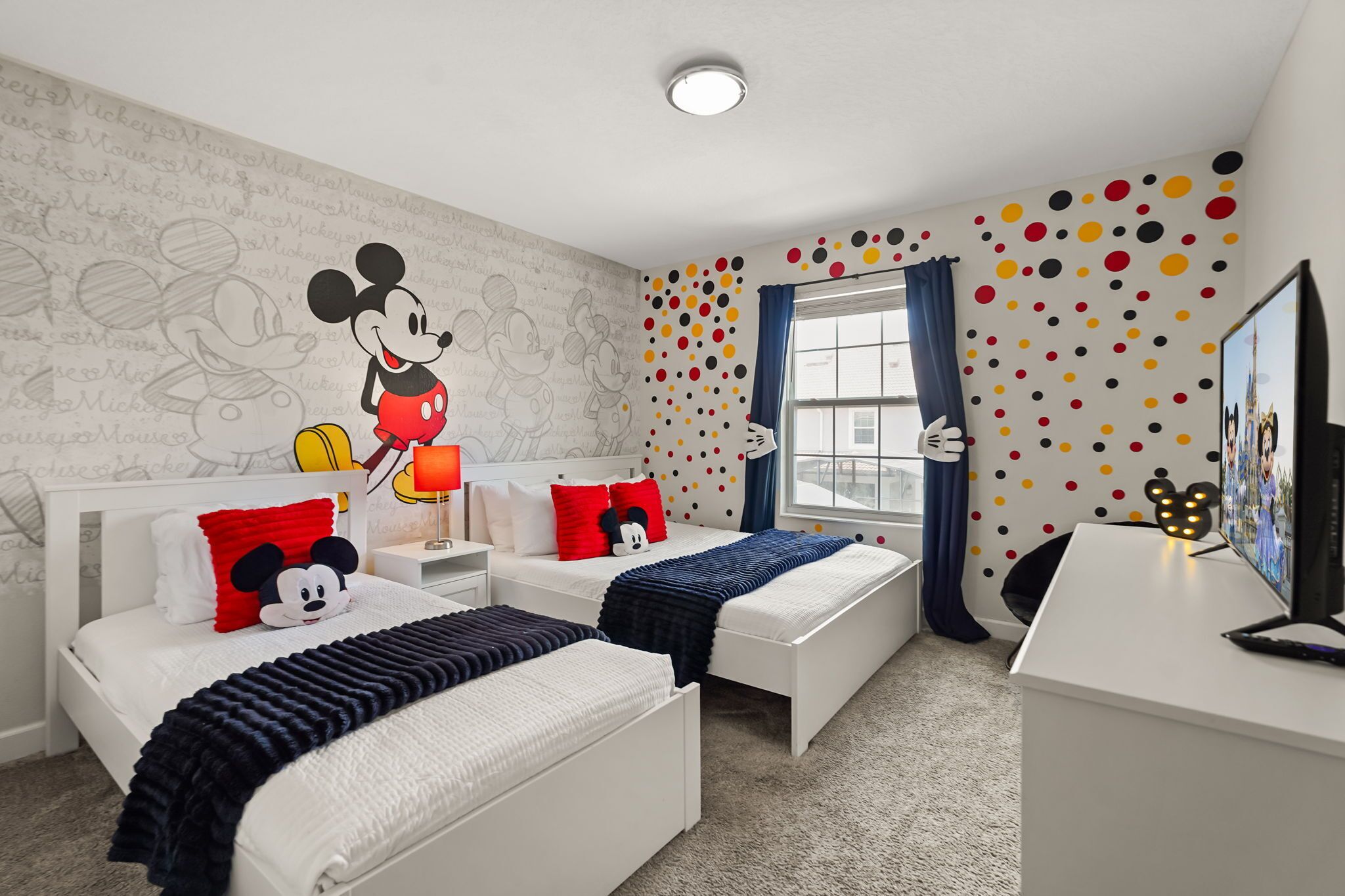 Habitación de hotel con decoración de Mickey Mouse, dos camas, un escritorio y una ventana con cortinas.