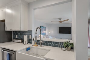 A modern kitchen with white cabinets, a built-in sink, a ceiling fan, and a window overlooking a bedroom. - Modern, Dog-Friendly Marsh Retreat (Mount Pleasant)