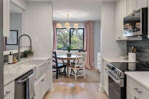 A modern kitchen with white cabinetry, a dining area with a wooden table and chairs, a built-in oven, and a large window with curtains. - Modern, Dog-Friendly Marsh Retreat (Mount Pleasant)