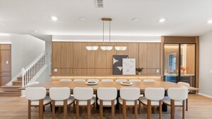 Un comedor moderno con una mesa de madera y sillas blancas, una escalera y una cocina visible a través de una puerta abierta.