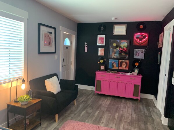 A room with a black sofa, a pink cabinet, and various framed items on the wall. - Fall Weekend Getaways -Groovy Vinyl -East Bay Inn (Panama City)