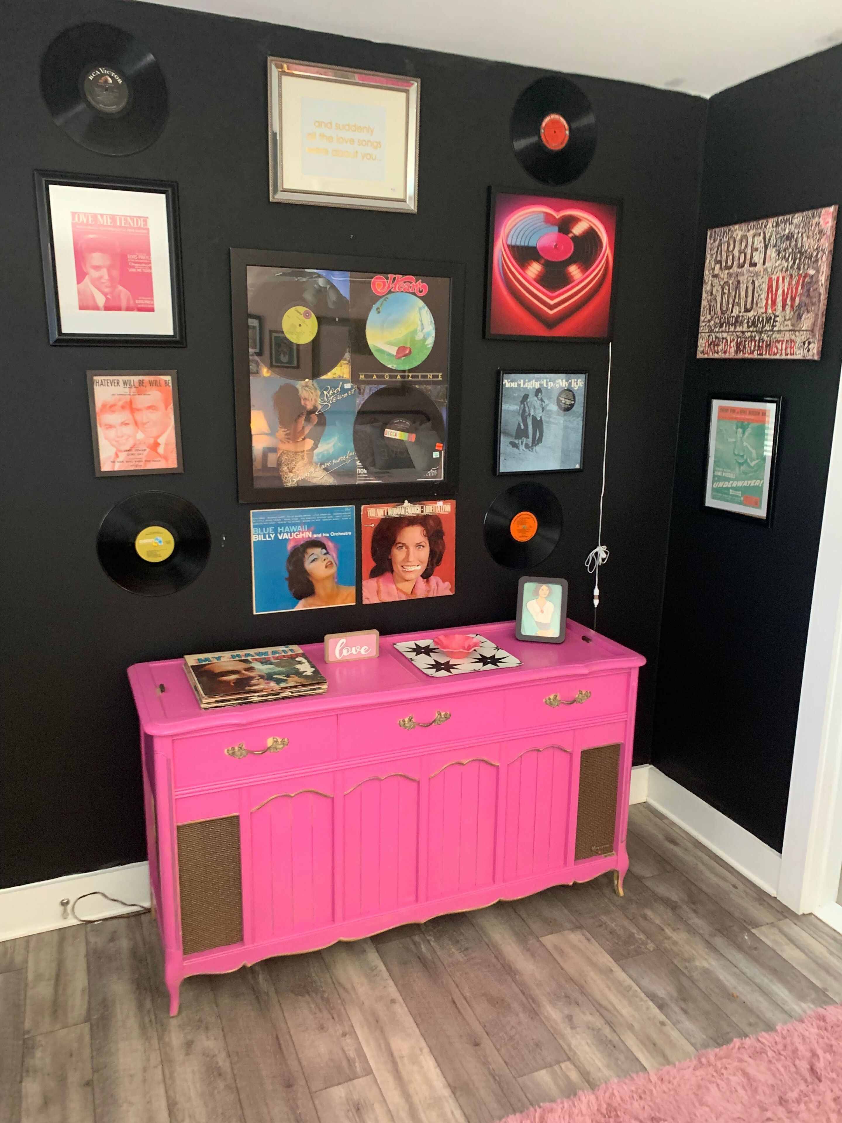 A bright pink vintage console cabinet rests against a black vinyl-themed gallery wall.