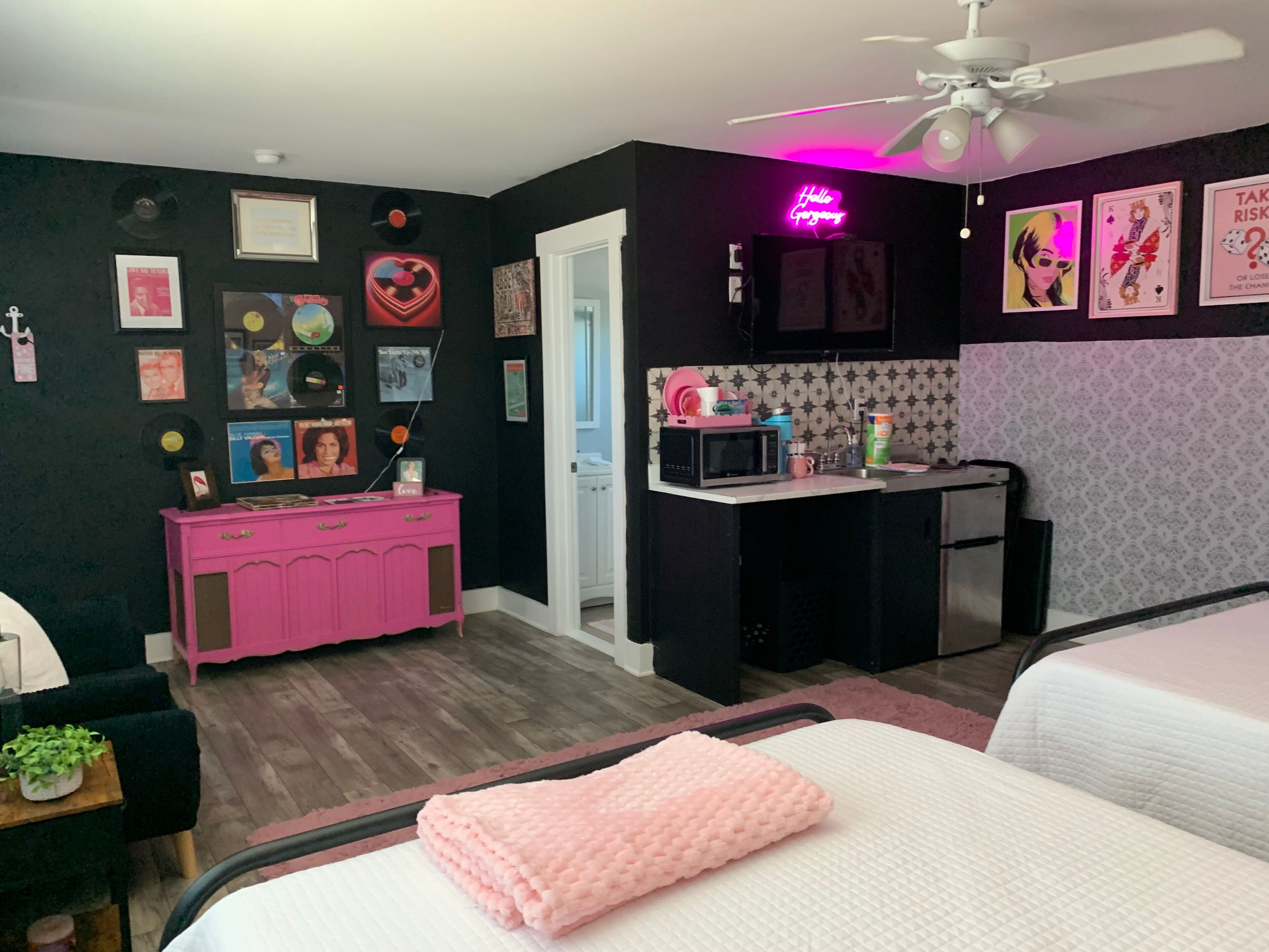 A room with a bed, a pink dresser, a microwave, a sink, and a ceiling fan.