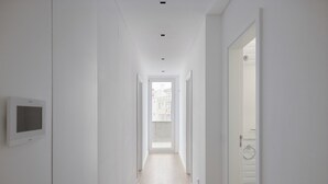 En smal hall med vita väggar, en takmonterad lampa, en dörr med en handdukskrok och ett fönster i änden som leder ut till en balkong.