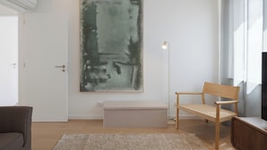 Ett rum med en stor, abstrakt målning, en bänk, en stol, en golvlampa och ett fönster med gardiner.