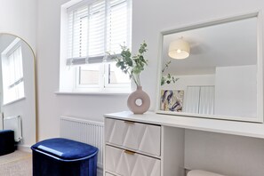 Ein Zimmer mit einem großen Spiegel, einer Kommode mit Schubladen, einem blauen Hocker und einem Fenster mit Jalousien.