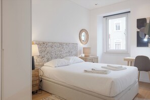 Et hotelværelse med en pænt redt seng, en sengetavlelampe, et lille bord, en stol og et vindue med udsigt til bygninger.