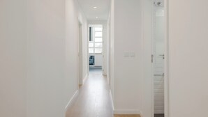 Passadís estret amb parets blanques i un terra de fusta, que condueix a una habitació amb finestra i porta.