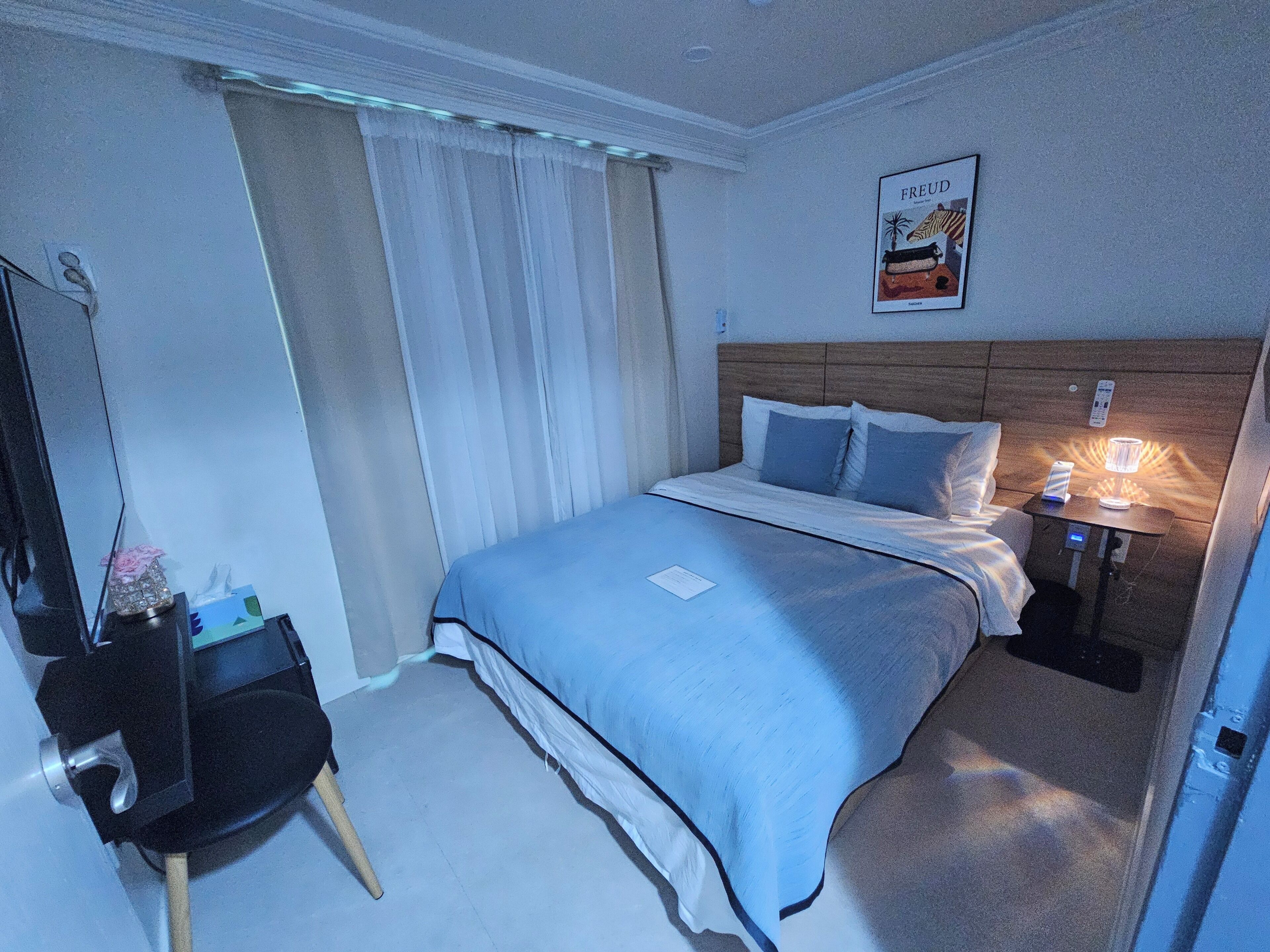 A hotel room with a bed, a nightstand with a lamp, a desk with a chair, and a poster on the wall.