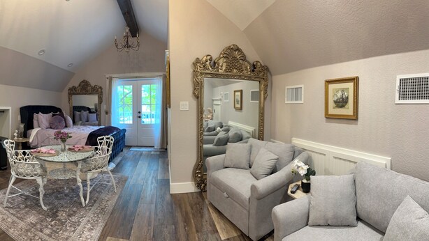 A compact living space with a kitchenette, a dining area with a table and chairs, a sofa, a large ornate mirror, and a view through a door to an outdoor area. - Saint Jo Wine Country Loft Near Blue Ostrich Winery (Saint Jo)