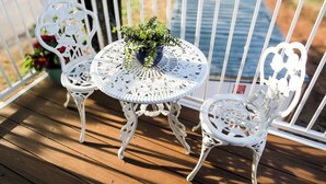A white metal table with a floral design, two white metal chairs, and a potted plant on a wooden deck.