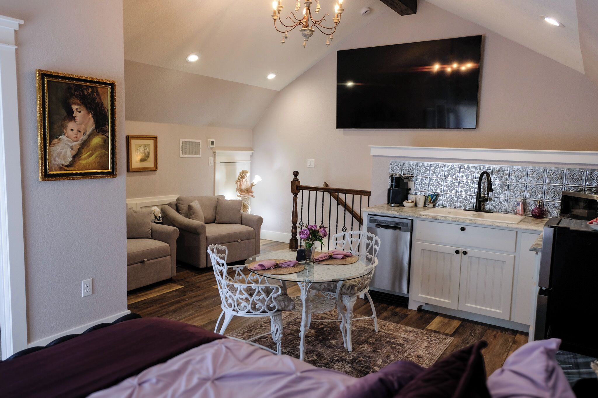 A cozy living area with a sofa, a dining table with chairs, a kitchen with a tiled backsplash, and a staircase.