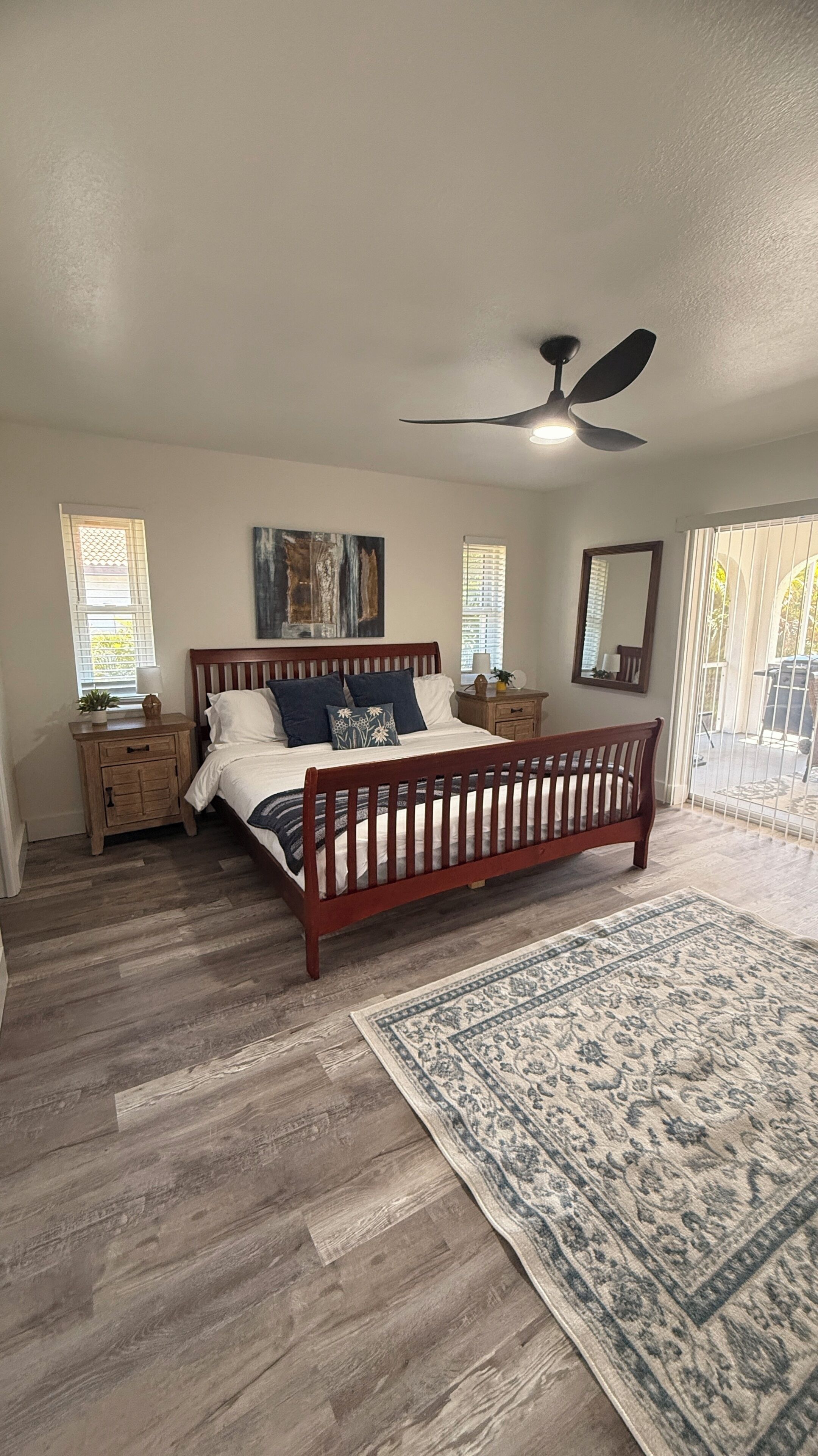 A bedroom with a wooden bed, a ceiling fan, a nightstand, a bedside table, a large rug, and a window with blinds.