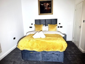Un lit recouvert d’une couverture jaune, avec une tête de lit sombre et un tableau encadré au-dessus.