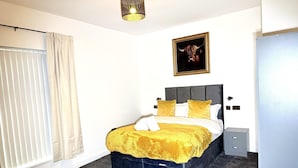 Een slaapkamer met een bed, een nachtkastje, een raam met gordijnen, een plafondlamp en een ingelijst schilderij aan de muur.