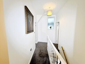 Un couloir orné d’un tableau encadré, d’une plante en pot, d’un escalier avec une rampe blanche et d’un luminaire.