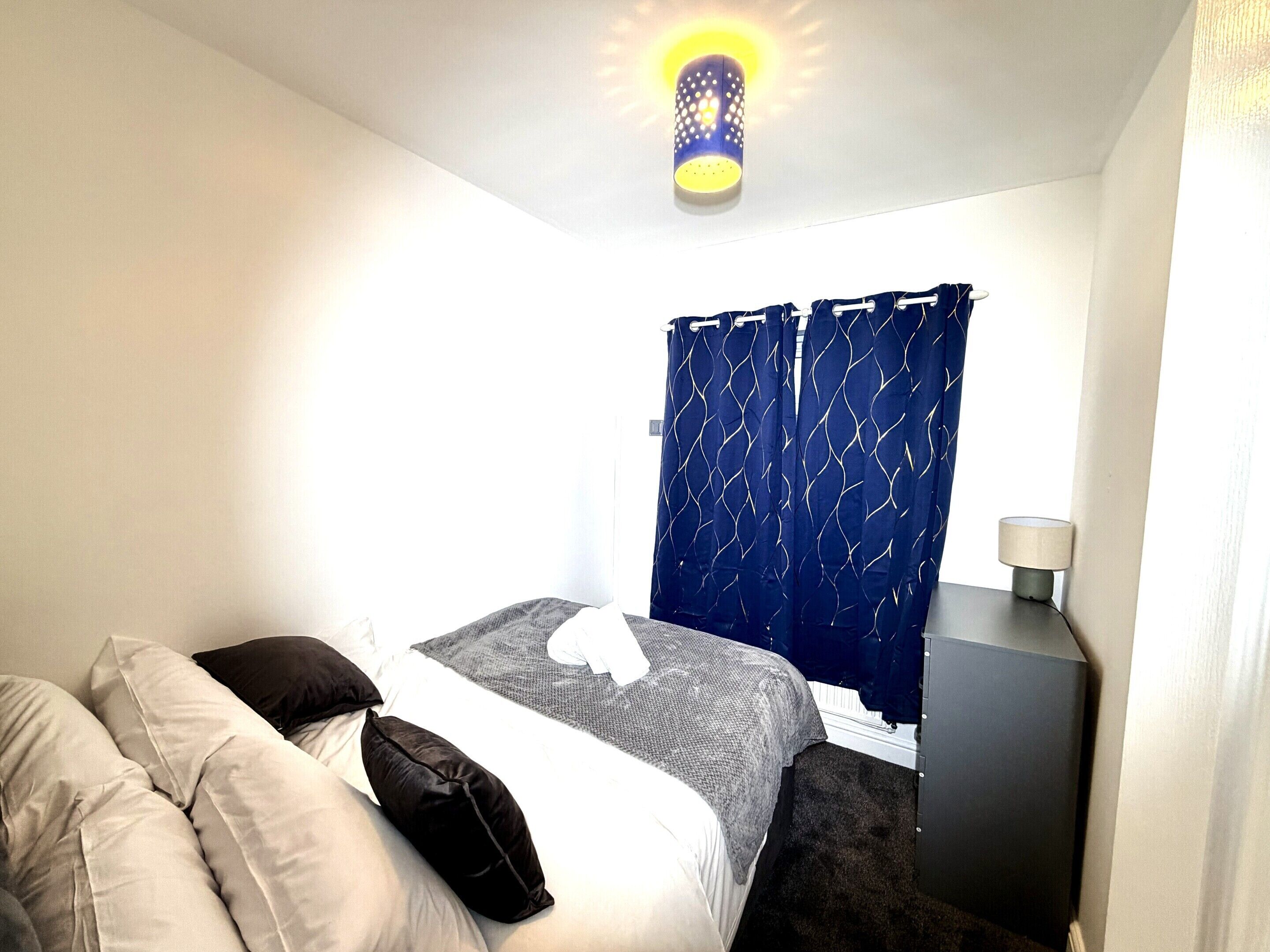 Een slaapkamer met een bed, kussens, een nachtkastje, een lamp en een raam met gordijnen.