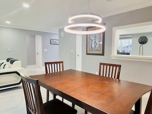 A dining area with a wooden table, four chairs, a modern pendant light, a framed artwork on the wall, and a window with a clock. - La B Villa (Cape Coral)