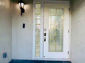 Una puerta blanca con un panel de vidrio y un diseño decorativo, una lámpara de pared y un pequeño cuadro eléctrico.
