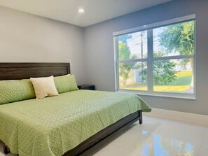A bedroom with a bed, a nightstand, a window with a view of greenery, and a white tiled floor. - La B Villa (Cape Coral)