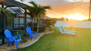 A patio with blue chairs and lounge chairs, a glass structure, and a grassy area. - Casa Azul/Tropical Paradise/Heated Pool/Private (Cape Coral)