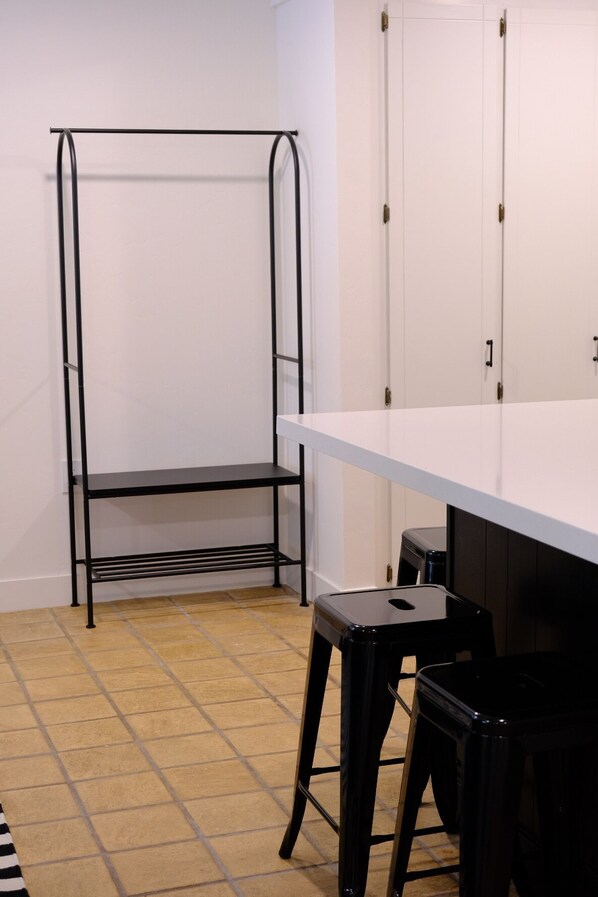 Una habitación con un perchero metálico, una mesa blanca y dos taburetes negros.