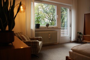 Ein Zimmer mit großem Fenster, Heizkörper, Stuhl, Nachttisch und Nachttischlampe.